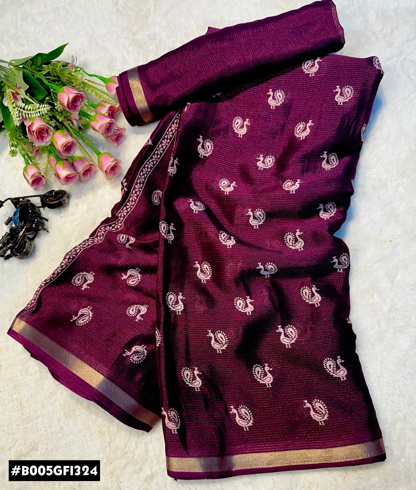 A folded deep purple/magenta saree with small white peacock motifs and a gold border, displayed on a light textured background with pink roses as props.